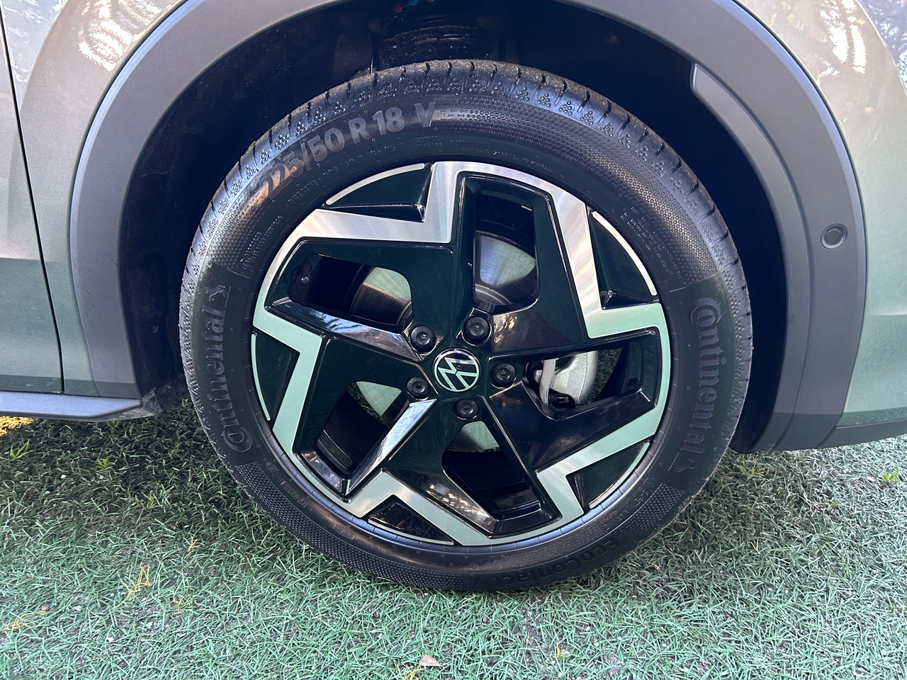 Close-up of a Volkswagen alloy wheel fitted with a Continental tyre on an SUV. The wheel features a black and silver multi-spoke design with the Volkswagen logo at the centre. The tyre sidewall displays the size marking "225/50 R18" and Continental branding. The wheel arch, grey bodywork, brake disc, and brake caliper are visible around the wheel, with the vehicle parked on a grass-covered surface in daylight.