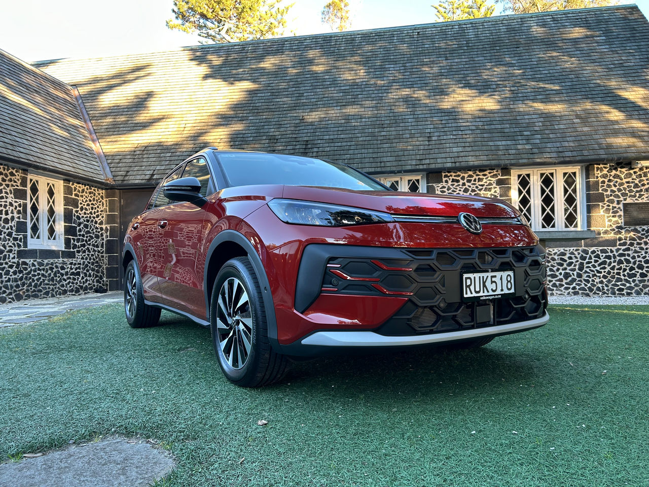 Front three-quarter view of a red Volkswagen SUV parked on a grassed area in front of a historic dark-stone building with a steep shingled roof and tall white-framed windows. The vehicle's grille, headlights, alloy wheels, and front license plate are clearly visible. Sunlight and tree shadows fall across the roof and the car, creating a bright outdoor setting.