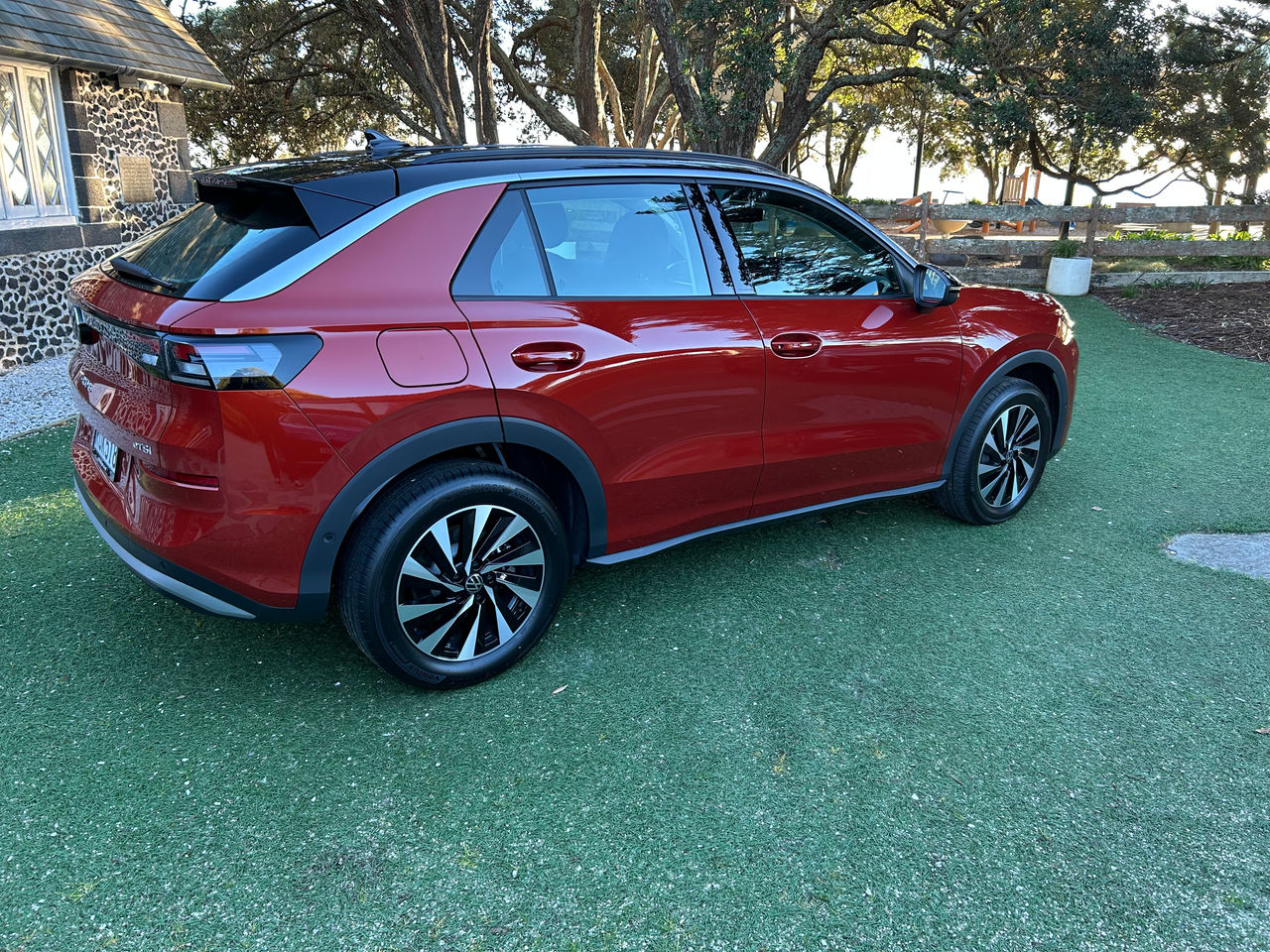 Rear three-quarter side view of a red Volkswagen T-Roc SUV parked on a grass-covered area beside a historic stone building. The vehicle features a contrasting black roof, black wheel arch cladding, tinted windows, roof spoiler, and multi-spoke alloy wheels. The sculpted bodywork, rear LED taillights, and sloping C-pillar design are visible from this angle. Mature trees, landscaped grounds, and timber fencing form the background, while natural daylight highlights the metallic red paint and exterior styling details. 