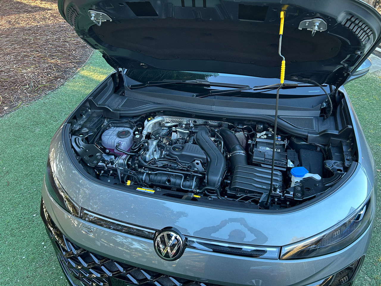 Overhead view of the engine bay of a silver Volkswagen T-Roc R-Line SUV with the bonnet open. Visible components include the engine cover, air intake system, battery, coolant reservoir containing pink fluid, washer fluid filler cap, and various hoses and wiring. The bonnet is supported by a prop rod, and the front Volkswagen badge, slim LED headlights, and upper grille are visible at the bottom of the frame. The vehicle is parked outdoors on a grass-covered surface in daylight.