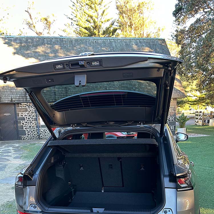 Rear view of a silver Volkswagen T-Roc with the tailgate raised, revealing the cargo area. The boot features a flat load floor, dark interior trim, a retractable parcel shelf, and split-fold rear seatbacks. The raised tailgate shows integrated lighting and latch mechanisms, while the vehicle is parked outdoors in front of a historic stone building and landscaped grounds. Natural daylight illuminates the luggage compartment and surrounding area.