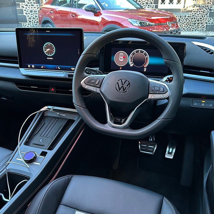 Driver’s-eye view of the interior of a Volkswagen T-Roc R-Line, showing a multifunction steering wheel with the Volkswagen logo, a fully digital instrument cluster, and a central touchscreen infotainment display. The centre console includes a wireless charging pad, rotary control, and storage compartment, while metal sport pedals are visible in the footwell. Black upholstery with contrast stitching and ambient lighting accents enhance the cabin. Through the windscreen, a red Volkswagen SUV and a stone building can be seen parked outside in daylight.