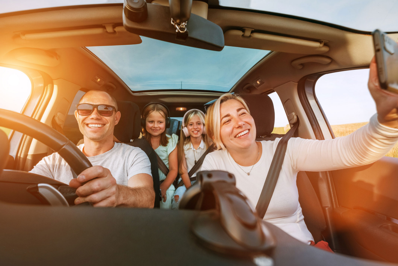 View from inside a car of four people on a road trip. One person is driving, another in the front passenger seat is holding up a smartphone for a photo, and two children are seated in the back. Warm sunlight streams through the windows and panoramic sunroof, illuminating the vehicle interior.