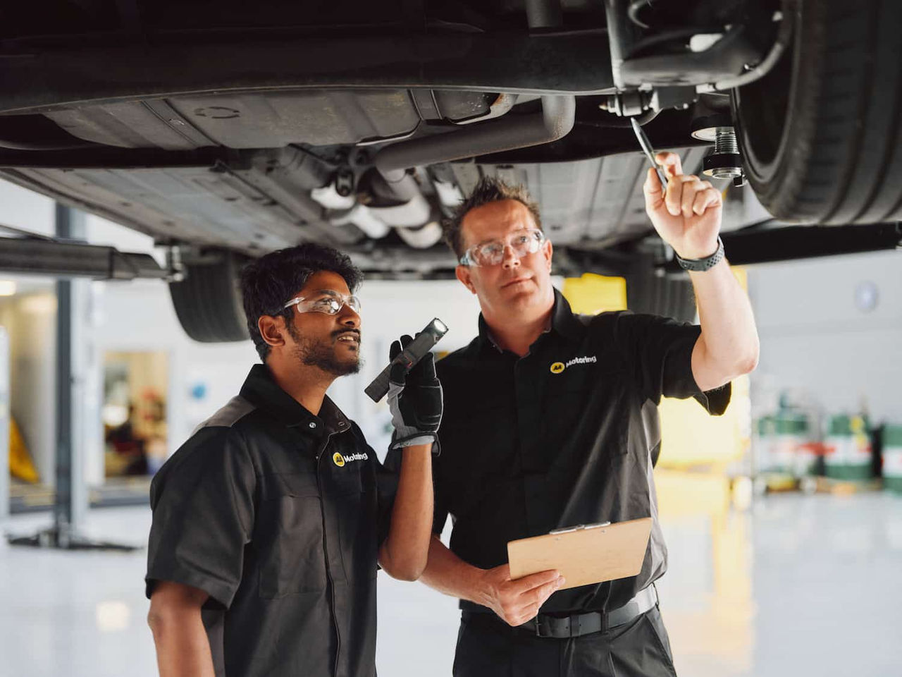 Two automotive technicians wearing safety glasses inspect the underside of a vehicle raised on a service hoist. One technician shines a flashlight toward vehicle components while the other points to an area near a wheel and holds a clipboard in a well-lit workshop.