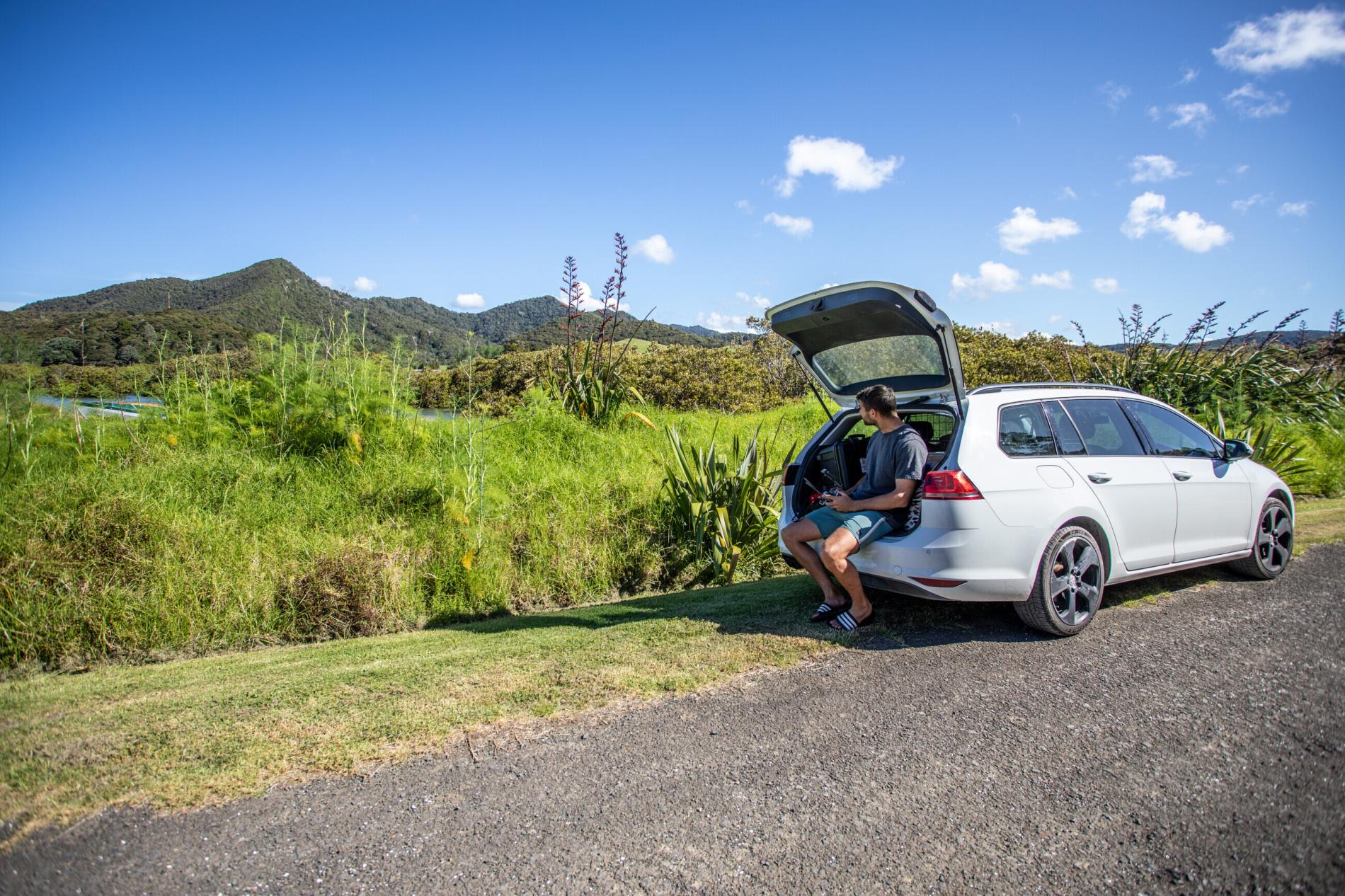 A man sitting in open car trunk, surrounded by greenery and hills