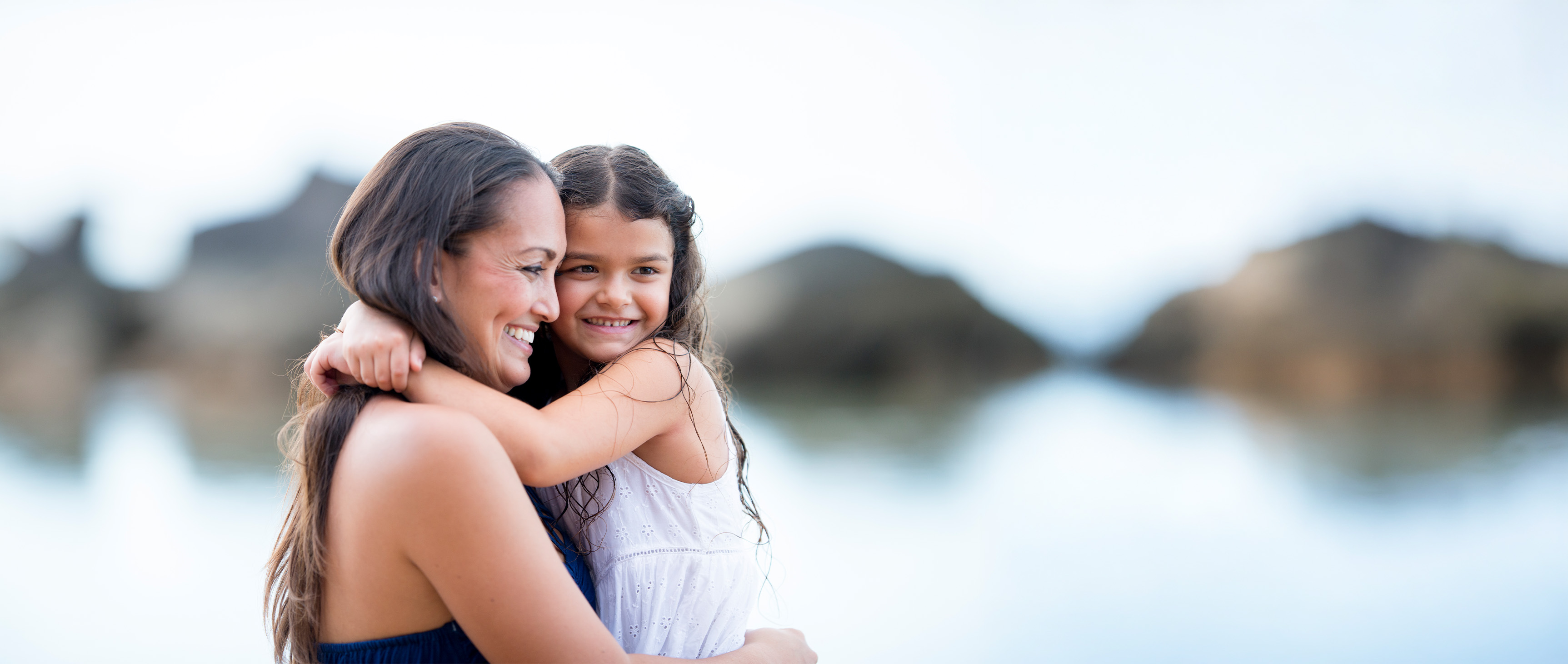 Woman holding her young daughter on a beach with water and land in the background