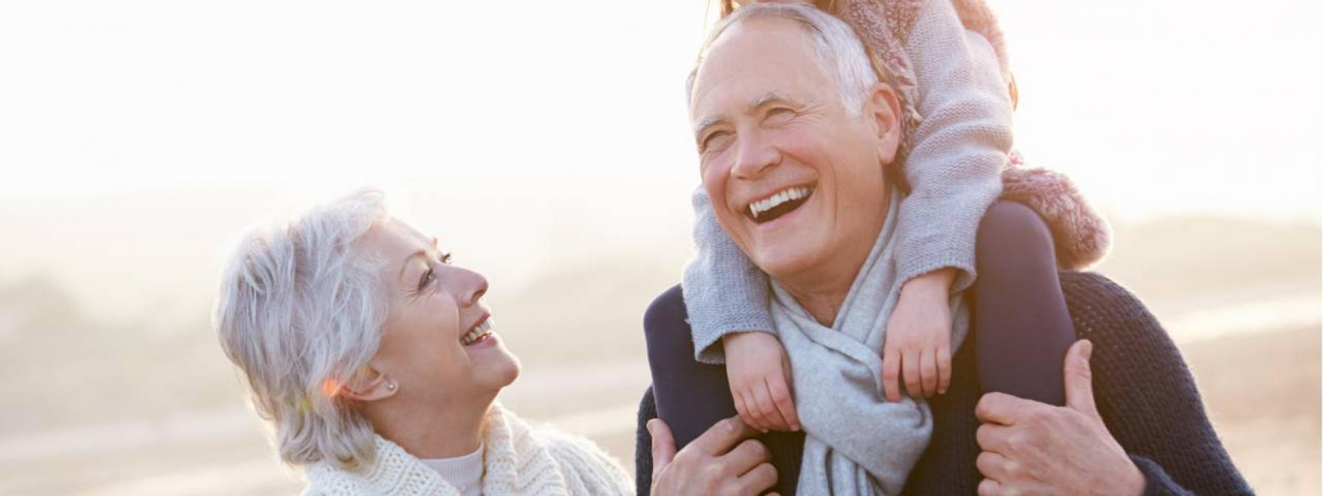 A grandmother and grandfather laughing while carrying his grandchild on his shoulders.