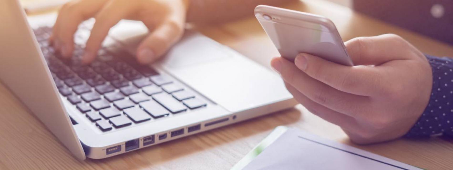 A hand holding a phone and the other typing on a laptop