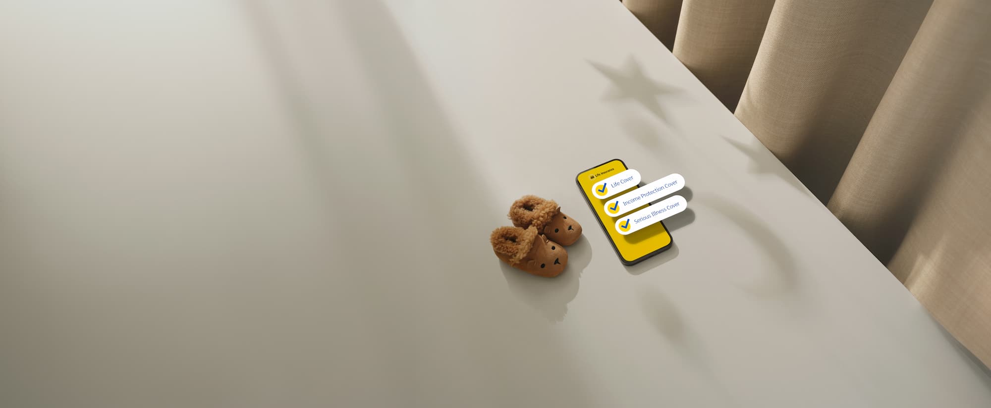 A pair of small brown fleece-lined baby booties sits beside a smartphone on a light-colored tabletop. The phone displays a yellow life insurance screen with three checked coverage options: “Life Cover,” “Income Protection Cover,” and “Serious Illness Cover.” Warm sunlight casts soft shadows across the surface, including star-shaped shadows near beige curtains in the background, creating a calm, family-focused atmosphere.