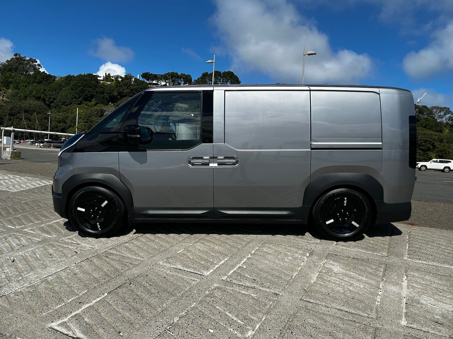 Side profile of a silver Kia PV5 electric cargo van parked outdoors on a concrete surface under a blue sky.