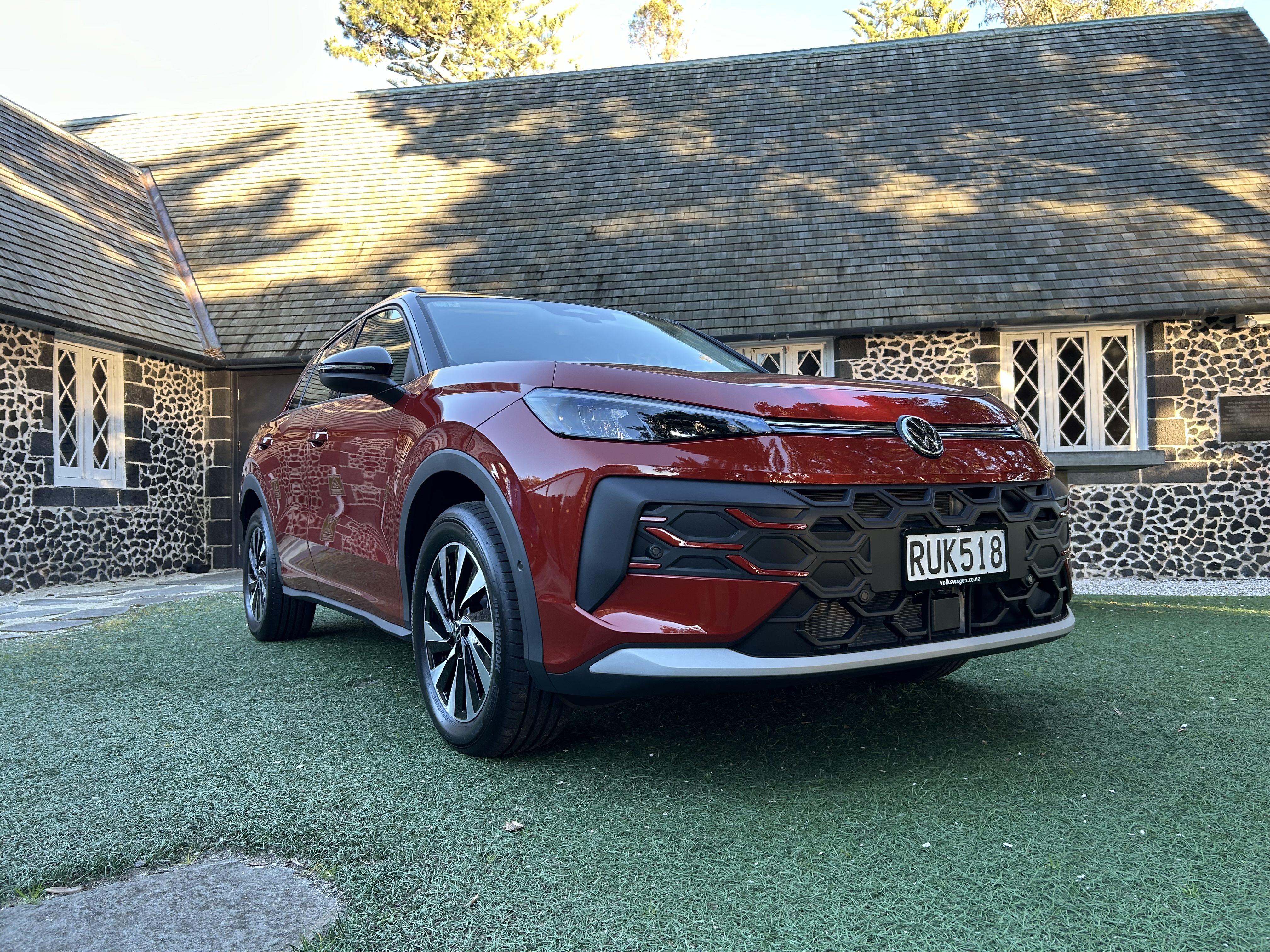 Front three-quarter view of a red Volkswagen SUV parked on a grassed area in front of a historic dark-stone building with a steep shingled roof and tall white-framed windows. The vehicle's grille, headlights, alloy wheels, and front license plate are clearly visible. Sunlight and tree shadows fall across the roof and the car, creating a bright outdoor setting.