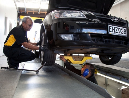 Two men inspect a tire on a lifted car for a Warrant of Fitness check in a workshop