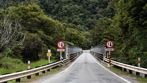 Narrow one-lane bridge surrounded by green forest and giveway signs on both sides
