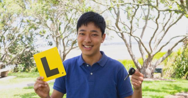 A young man smiling at the camera while holding a learners license plate and car keys