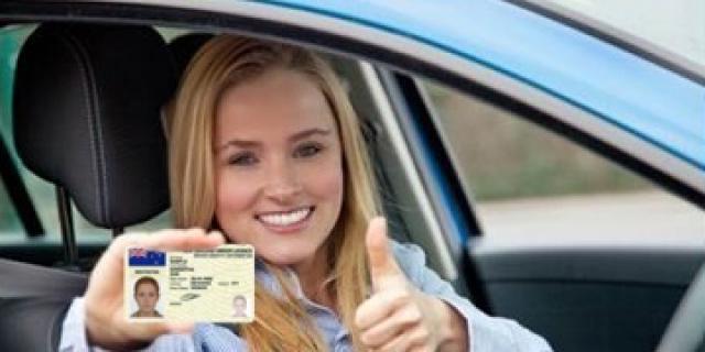 A young woman inside a vehicle smiling while looking at the camera holding her driver license