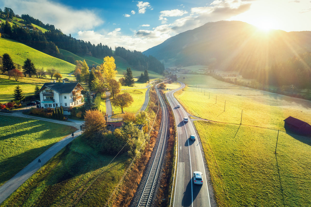 Aerial view of the road in mountain valley at sunset in spring in Dolomites, Italy. Top view of cars on asphalt roadway, house, railroad, hills with green meadows, blue sky, trees, buildings. Highway