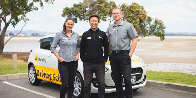 Three people standing next to a car with an AA Driving School logo, with a car in the background for training