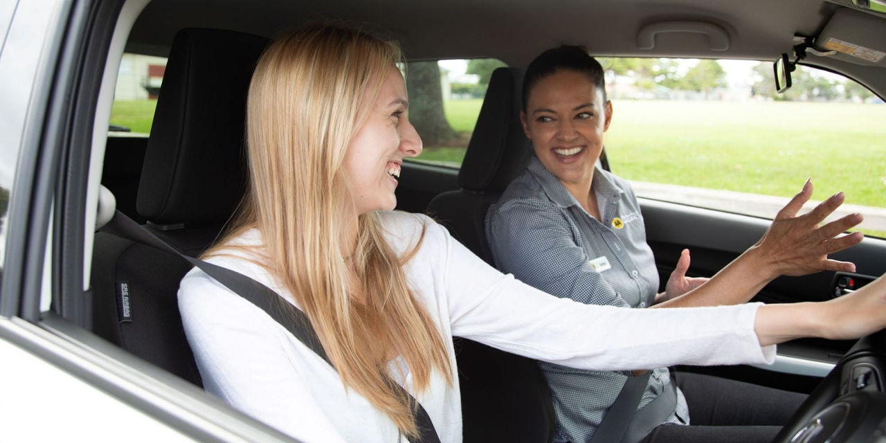 Young woman in the front seat of her car taking feedback from an AA Driving Instructor in the passenger seat.