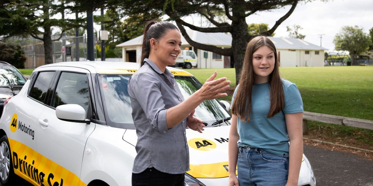 A woman driving instructor talks to a learner driver while standing beside a parked car
