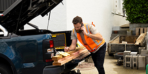 Man in an orange fluoro vest writing on planks of wood that are being loaded into the back of a ute.