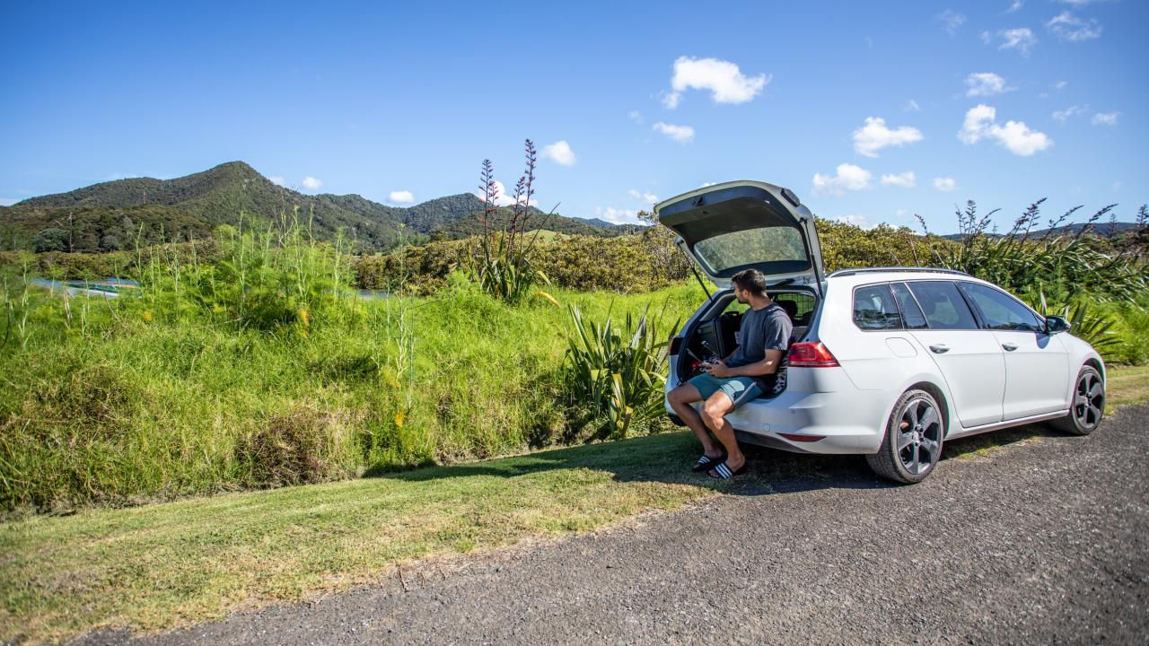 A man sitting in open car trunk, surrounded by greenery and hills
