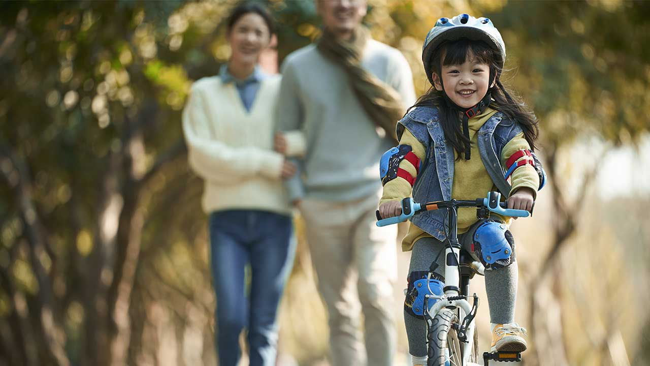 Young girl riding her bike as her parents watch from the background.