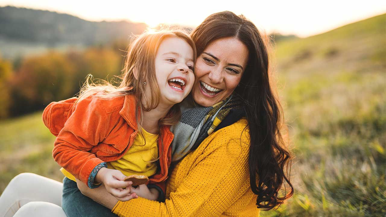 A mother and her young daughter laughing together on a hillside.