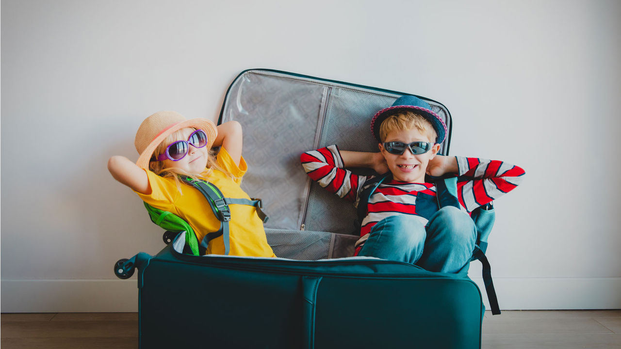 Two children wearing traveling gear sitting inside a suitcase together