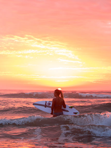A person holding a surfboard is standing in shallow waves looking at a sunset on the horizon.