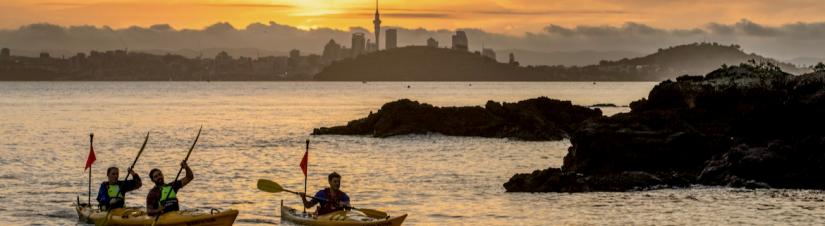 Three people kayaking with the Auckland city skyline and sunset in background