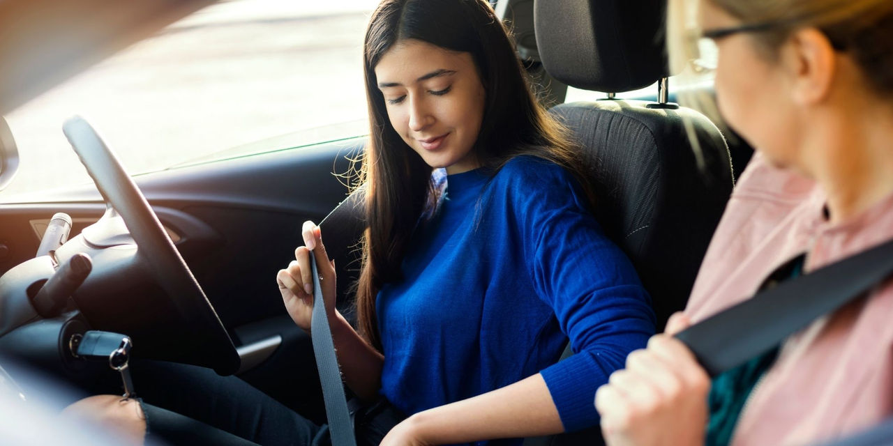 Inside a parked vehicle, a person seated in the driver's seat is fastening a seatbelt across the body before driving. Another person seated in the front passenger seat is also wearing a seatbelt. The vehicle interior, steering wheel, front seats, and dashboard are visible, with daylight entering through the windows. The image highlights safe driving practices and the importance of wearing a seatbelt.
