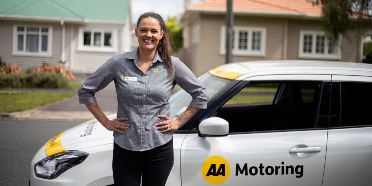 A person wearing an AA Motoring branded shirt stands beside a white AA Motoring vehicle parked in a residential street. The vehicle displays the AA Motoring logo on the side and learner driver markings in yellow and white. Houses and greenery are visible in the background.