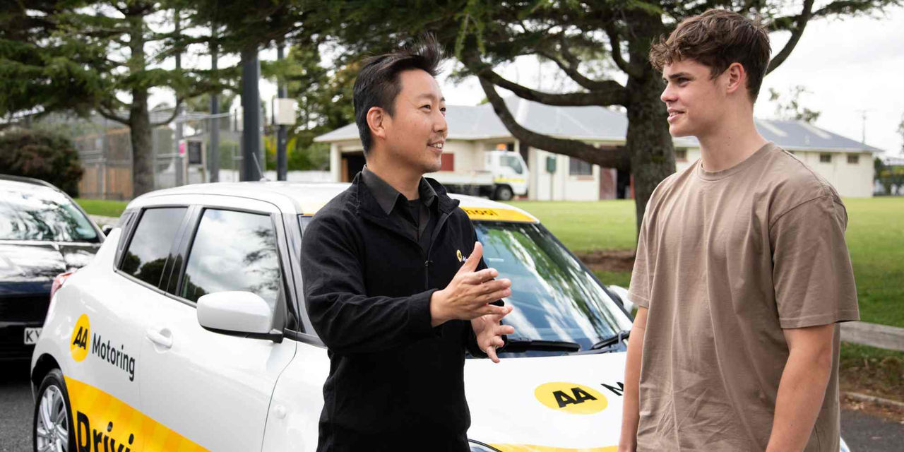 Two people stand beside a white AA Motoring learner driver vehicle in an outdoor parking area. One person is gesturing while speaking, and the other is listening. The vehicle displays AA Motoring branding and learner driver signage. Trees, parked vehicles, and buildings are visible in the background, illustrating a driver training or driving lesson setting.