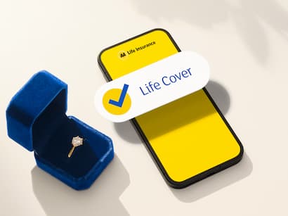 An open blue velvet ring box containing a diamond engagement ring sits beside a smartphone on a light-colored surface. The phone displays a bright yellow life insurance screen with a large checkmark and the text “Life Cover.” Soft natural light casts gentle shadows across the scene, including the silhouette of flowers in the background, creating a warm and elegant atmosphere associated with significant life milestones and future planning.