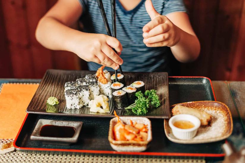 A focal image of a child using chopsticks to eat from a plate of Japanese sushi and other assorted foods 
