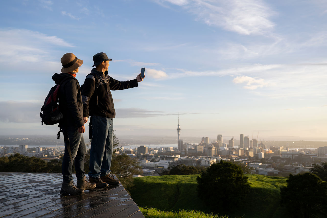 Couple taking photo with smartphone on Mt Eden summit at sunrise. Skytower in the background. Selective focus on people in foreground. Auckland. 