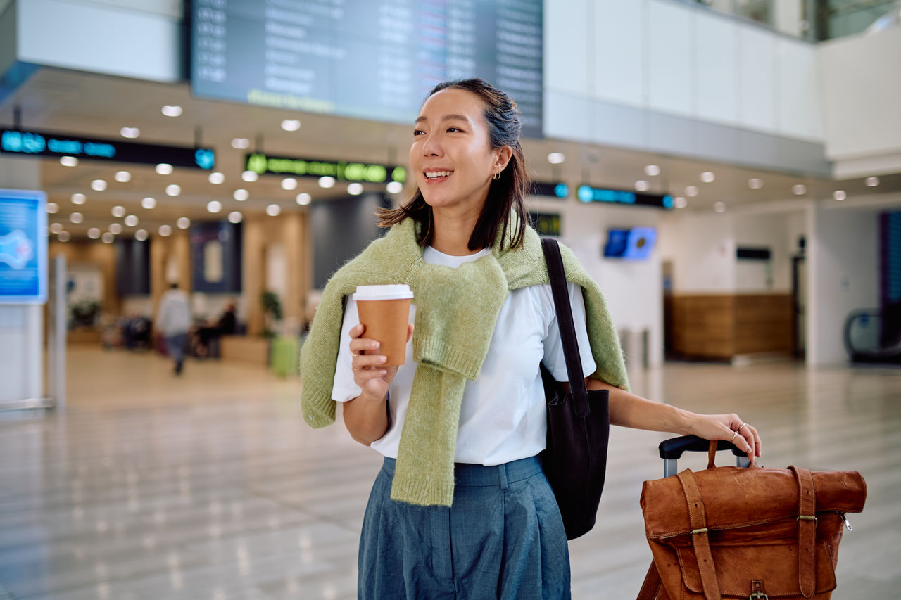 Side view young woman sits in a chair next to a yellow suitcase, drinking coffee and gazing out the window while waiting for her flight in an airport waiting lounge.