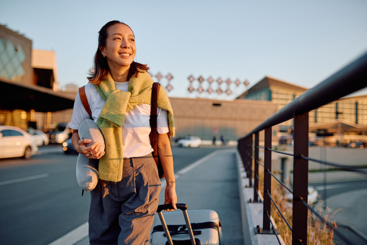 Happy young woman traveler walking with suitcase and neck pillow at an airport at sunset
