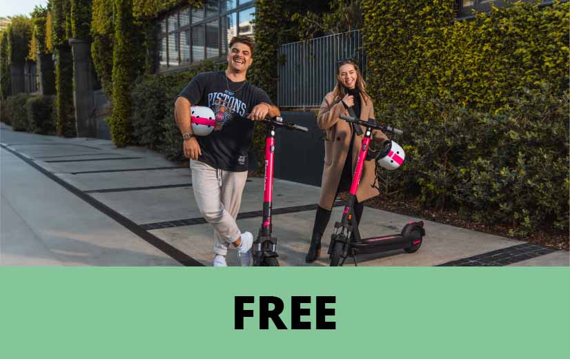 Two people standing with pink-and-black electric scooters on a paved urban walkway beside a building covered in greenery, each holding a white helmet. A green banner at the bottom of the image displays the word “FREE” in large black letters.
