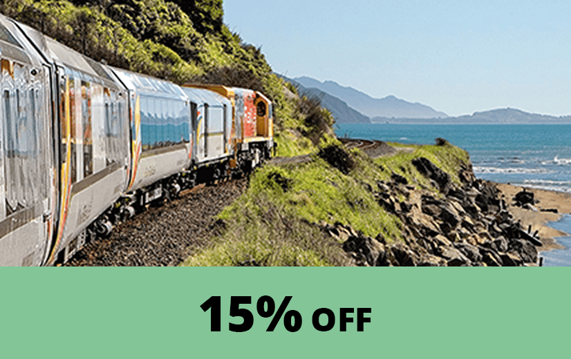 A scenic passenger train travels along a coastal railway track beside the ocean, with green hills and rocky shoreline in the background. A green banner at the bottom displays the text ‘15% OFF