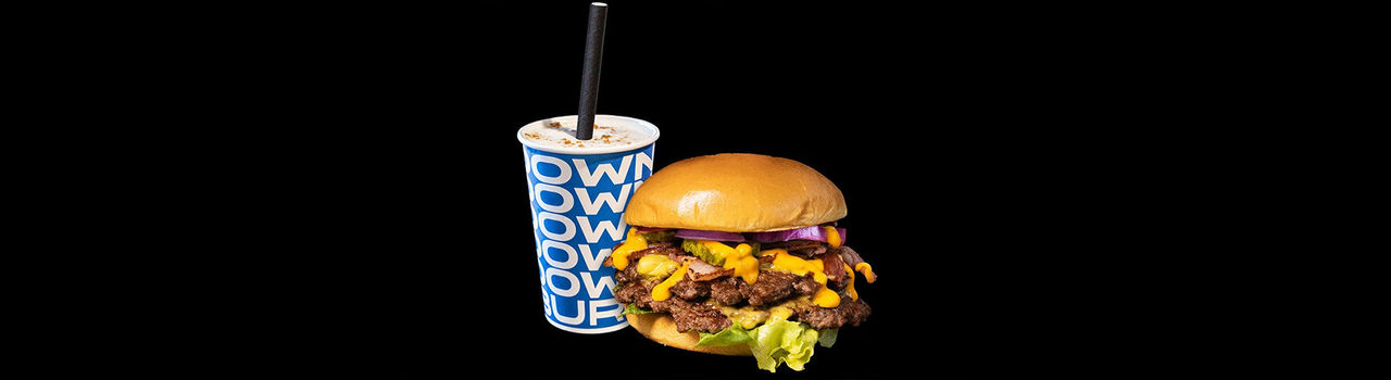 A large cheeseburger topped with melted cheese, bacon, lettuce, pickles, and red onion sits beside a thickshake in a blue branded cup with a black straw. The food is displayed against a solid black background, creating a high-contrast promotional-style image.