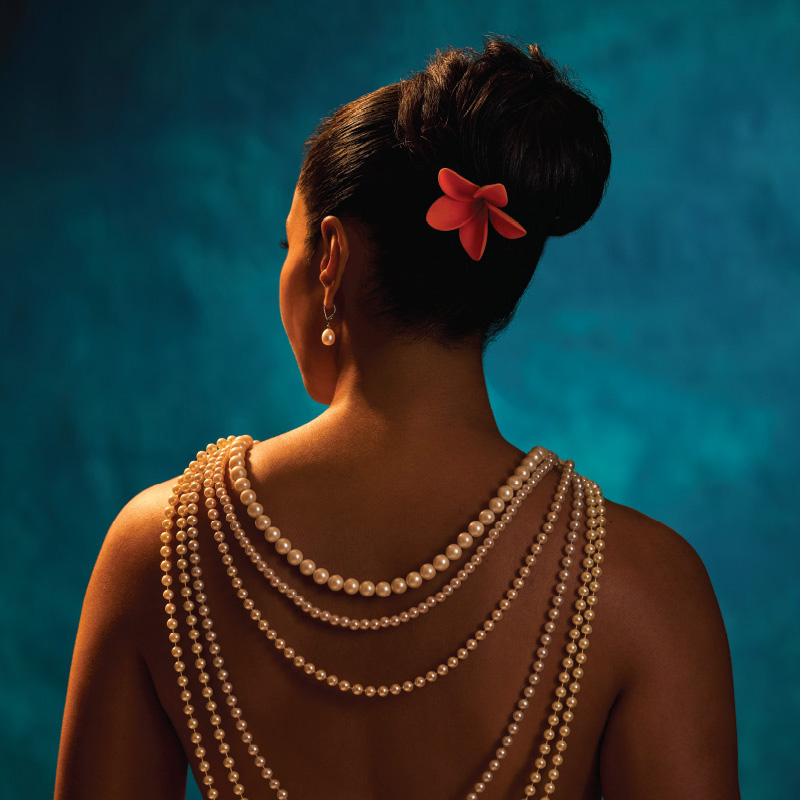 Person shown from behind with dark hair pinned up and decorated with a pink flower. Multiple strands of white pearls drape across the bare back and shoulders, with a pearl earring visible. The subject is illuminated by warm lighting against a textured blue background.