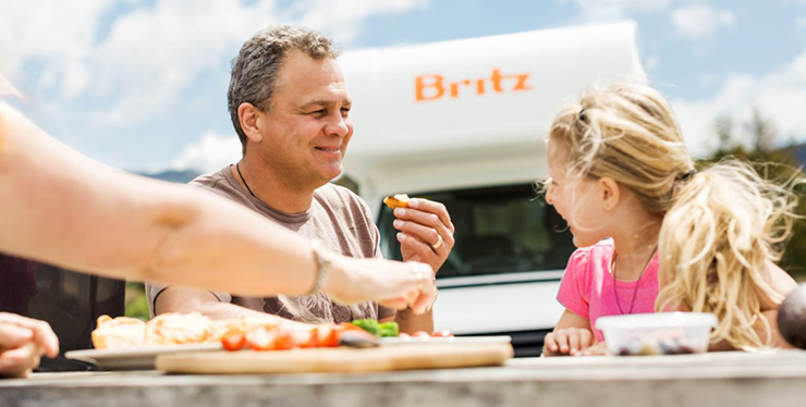A father and daughter sharing food on a picnic bench with an Apollo campervan in the foreground.