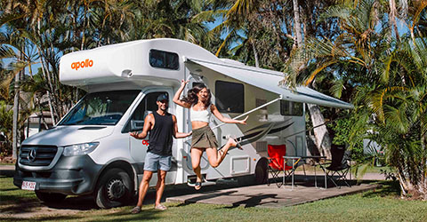 Young couple, woman jumping in the air with her partner behind her next to an Apollo campervan in the background.