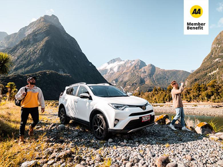 Two men standing either side of a parked car. They are admiring a scenic view between a river bed and mountains.